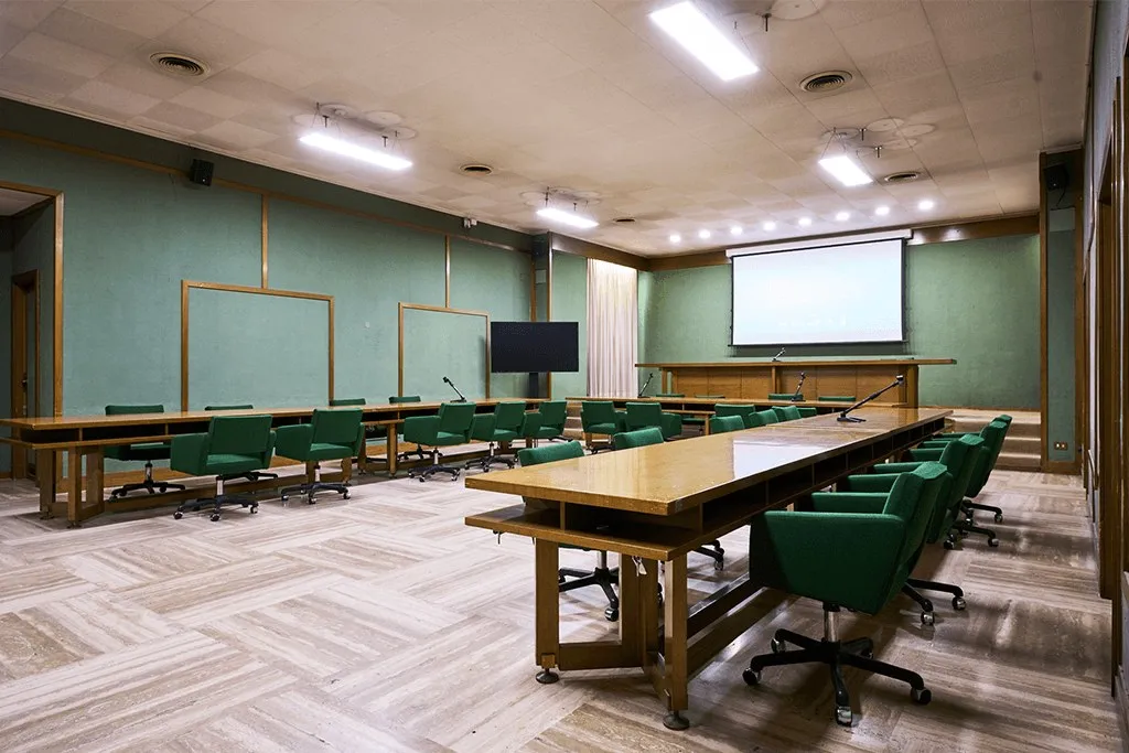 Sala riunioni con tavoli a ferro di cavallo, sedute verdi e schermo di proiezione